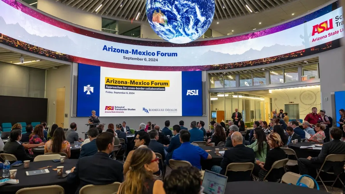 Woman speaking behind a lectern in front of a large screen that reads "Arizona-Mexico Forum" as a large audience looks on. Woman speaking behind a lectern in front of a large screen that reads "Arizona-Mexico Forum" as a large audience looks on.