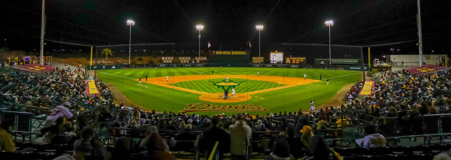 An image of the baseball field during a game in the evening with a crowd of fans in the foreground.