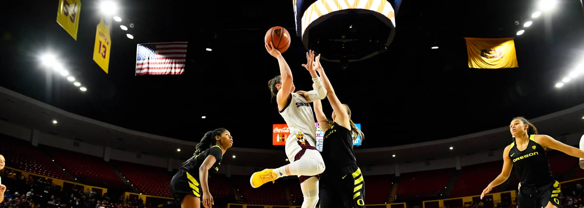 A female ASU basketball player jumps in the air to make a basketball hoop shot while the opposing team jumps up to stop her.