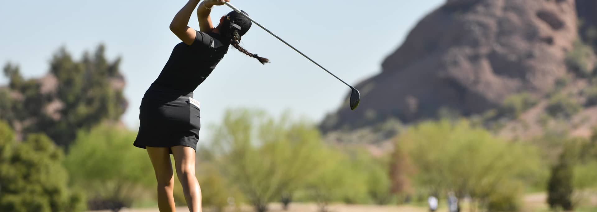 A female student golf player is shown in-action just after hitting a golf ball.