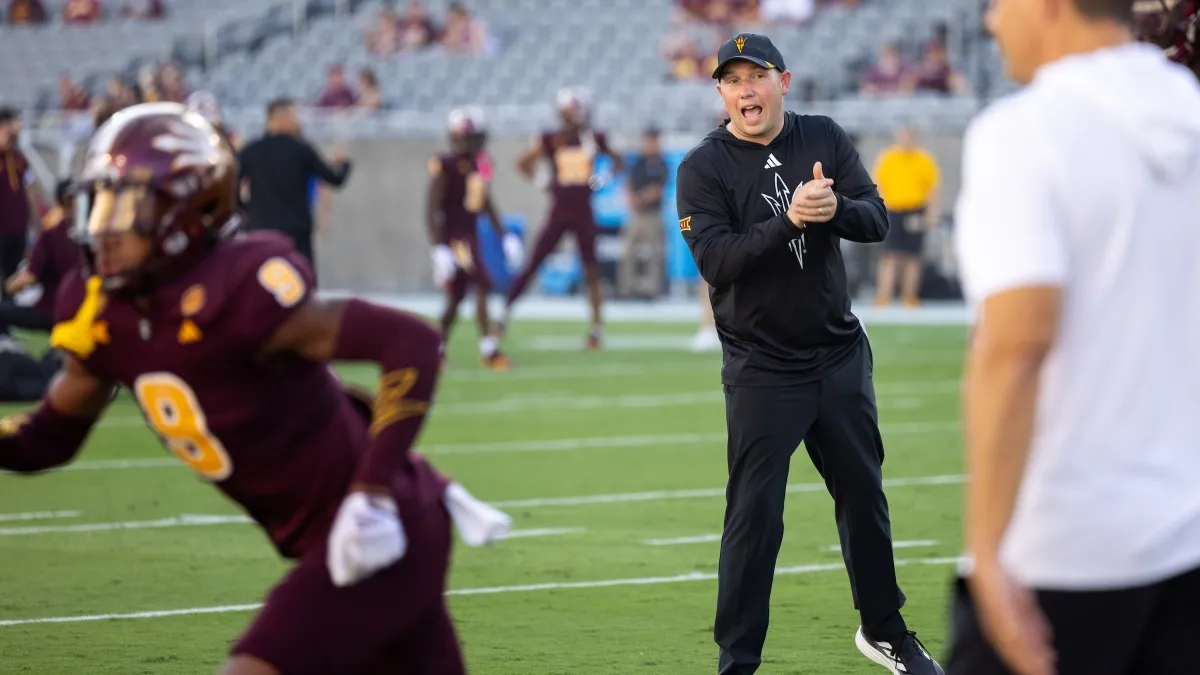 A man in a black tracksuit shouts to football players on the field as he coaches a game