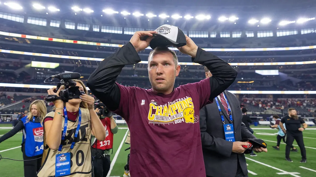 Man on a football field wearing a maroon shirt that reads &#039;champions&#039; as he puts on a hat that says the same.