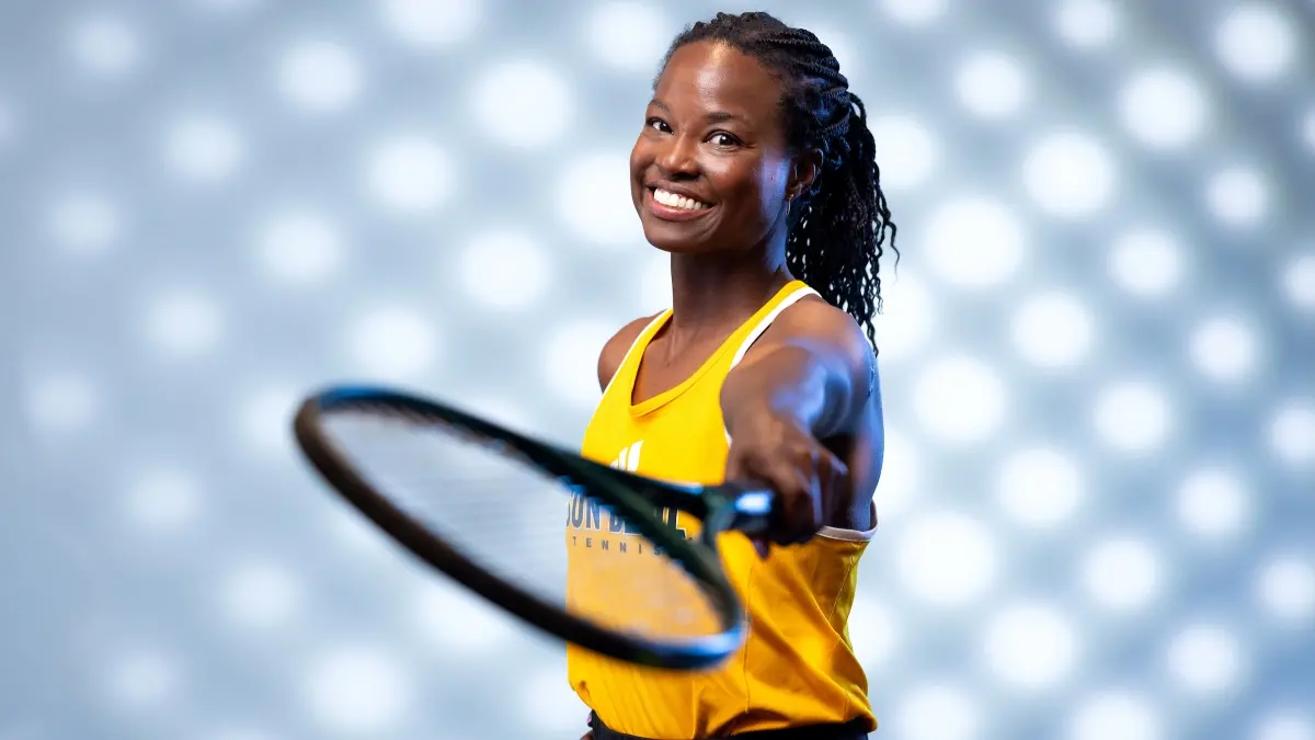 Portrait of a black woman with hair pulled into ponytail wearing a gold tank top and holding a tennis racket toward the camera