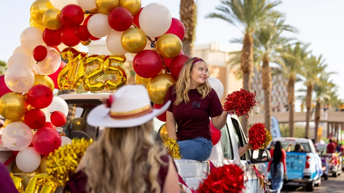 A car decorated in maroon, gold and white balloons with a young woman riding in the back drives in a Homecoming parade
