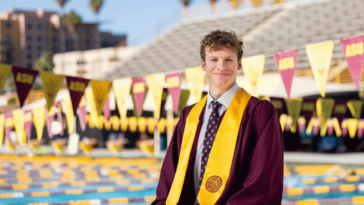 A young man in graduation regalia sits by the ASU swimming pool