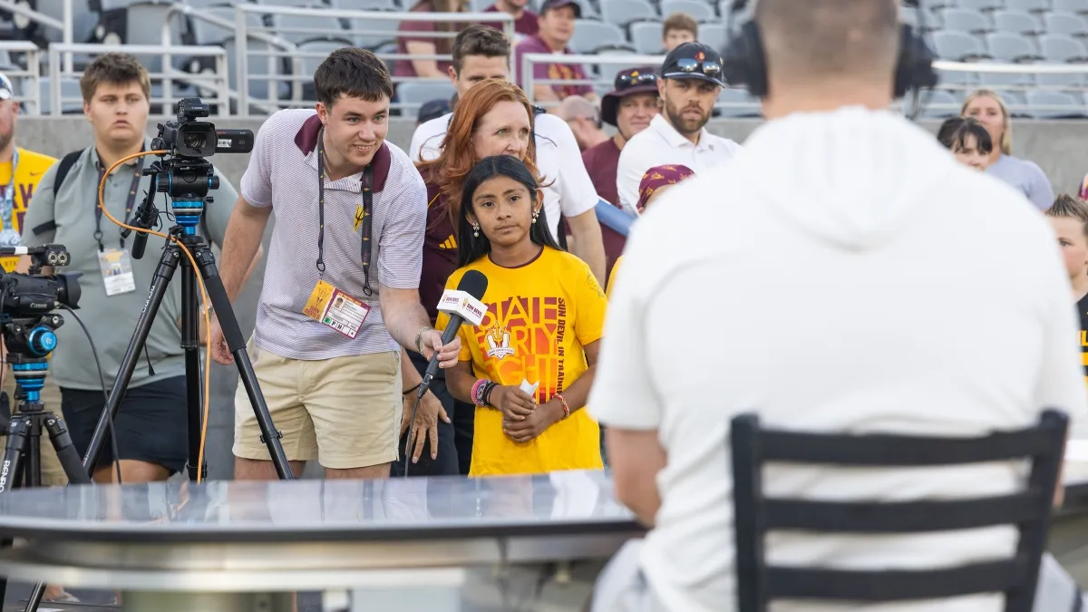 A young girl asks a coach a question while pretending to be a reporter at a press conference