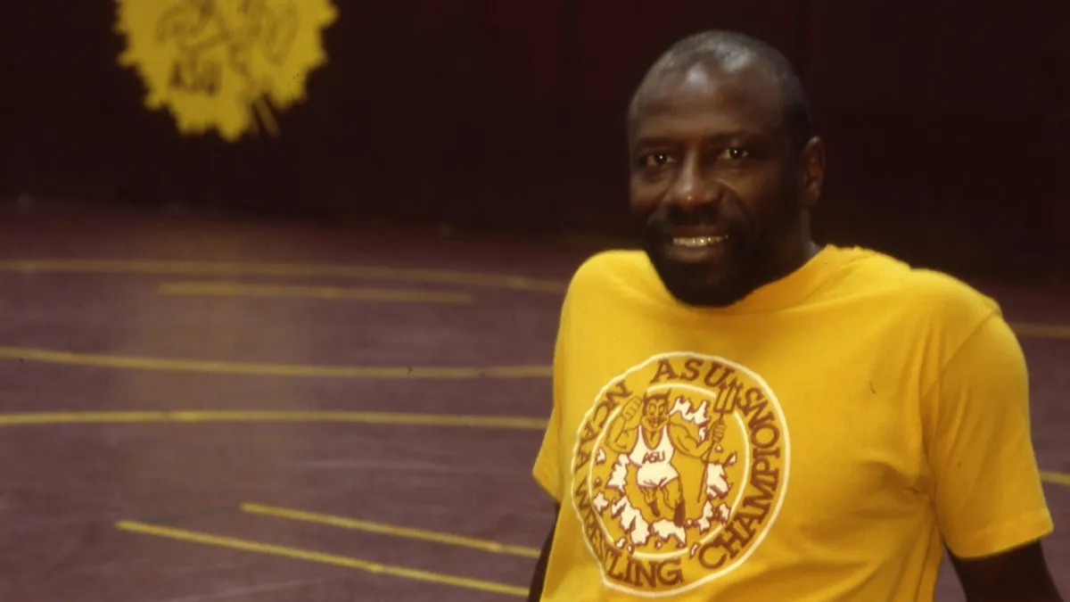 Portrait of a Black male wrestling coach wearing a gold ASU wrestling champions tshirt and maroon sweatpants