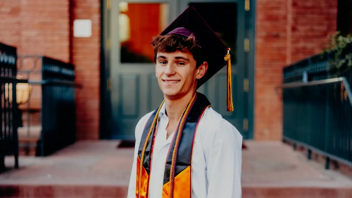 A young man stands smiling in a graduation cap, draped in graduation cords and vestments