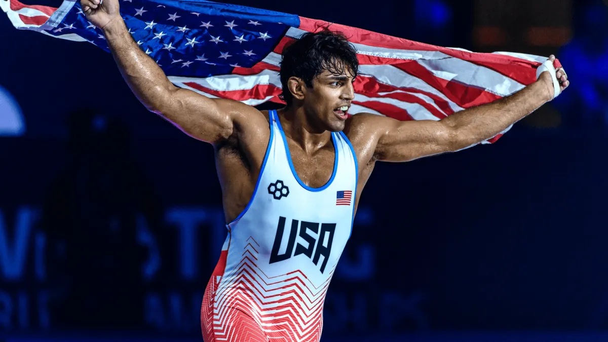 A male wrestler wearing a USA uniform runs holding an American flag above his head