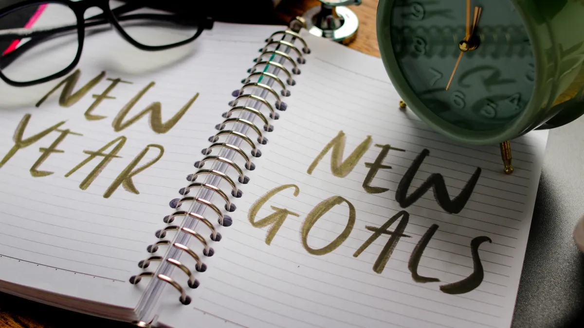 Stock photo with a notebook opened with the words New Year New Goals written in marker and a clock on top