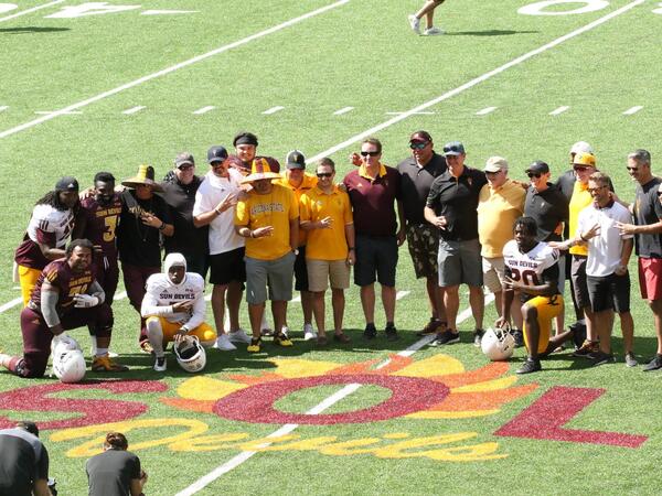 Group photo of ASU players and families on a football field with SOL Devils painted on the grass in front of them.